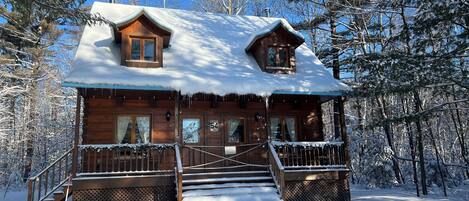A wooden cabin with a snow-covered roof, a porch with railings, and a staircase leading to the entrance.