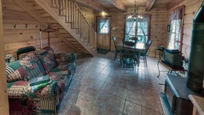 Interior acogedor de una cabaña con una escalera de madera, una zona de comedor con mesa y sillas, un sofá con una manta estampada y un estufa de leña.