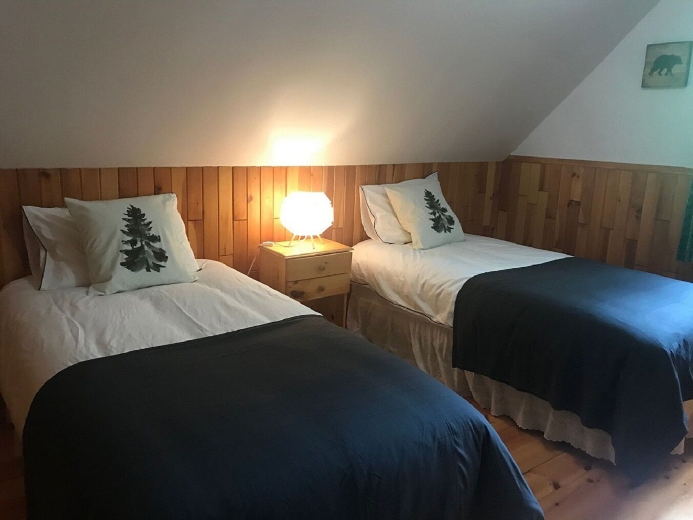 A room with two beds, each with a pillow featuring a tree design. There is a bedside table with a lamp between the beds.