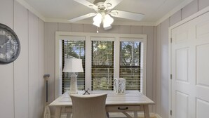 Ein Zimmer mit Deckenventilator, Tisch mit Stuhl, Lampe und einem Fenster mit Jalousien.