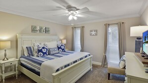 Ein Schlafzimmer mit Bett, Nachttisch, Schreibtisch, Stuhl, Deckenventilator und einem Fenster mit Vorhängen.