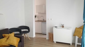 Ein modernes Wohnzimmer mit einer Couch, Sesseln, einem kleinen weißen Tisch und einer Küche im Hintergrund.