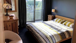 A bedroom with a bed, a desk with a lamp, a chair, a window with curtains, and a view of greenery outside. - Luxury Ouseburn flat with city & river views (Tyne and Wear)