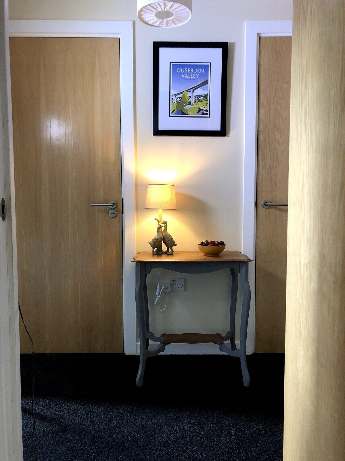Un couloir avec deux portes en bois, une petite table avec une lampe, un tableau encadré représentant la vallée d’Ouseburn et une prise électrique au mur.