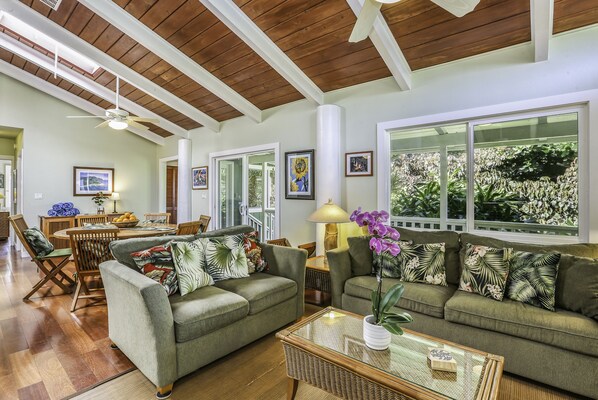 A spacious living room with a high ceiling, wooden beams, and large windows offering a view of greenery. - Haena Place, Walk to Beach Tropical Home TVNC#4232 (Haena)