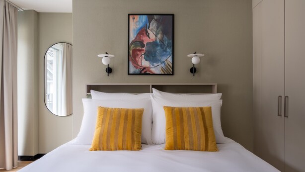 A hotel room with a large bed, two bedside lamps, a painting above the bed, and a wardrobe. - Numa Zurich Craft (Zürich)