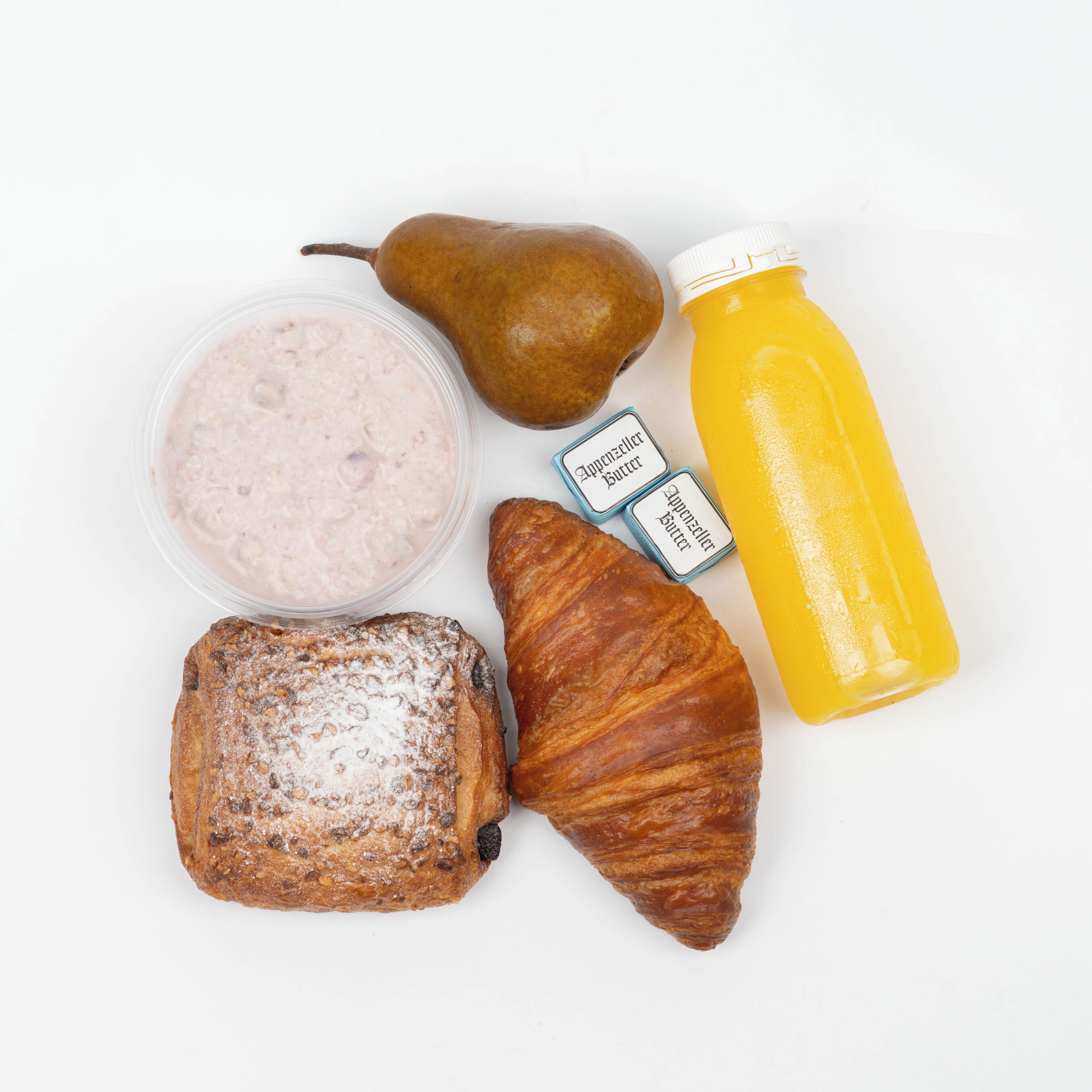 A breakfast setting with a pear, a croissant, a slice of bread, a container of yogurt, two packets of butter, and a bottle of orange juice.