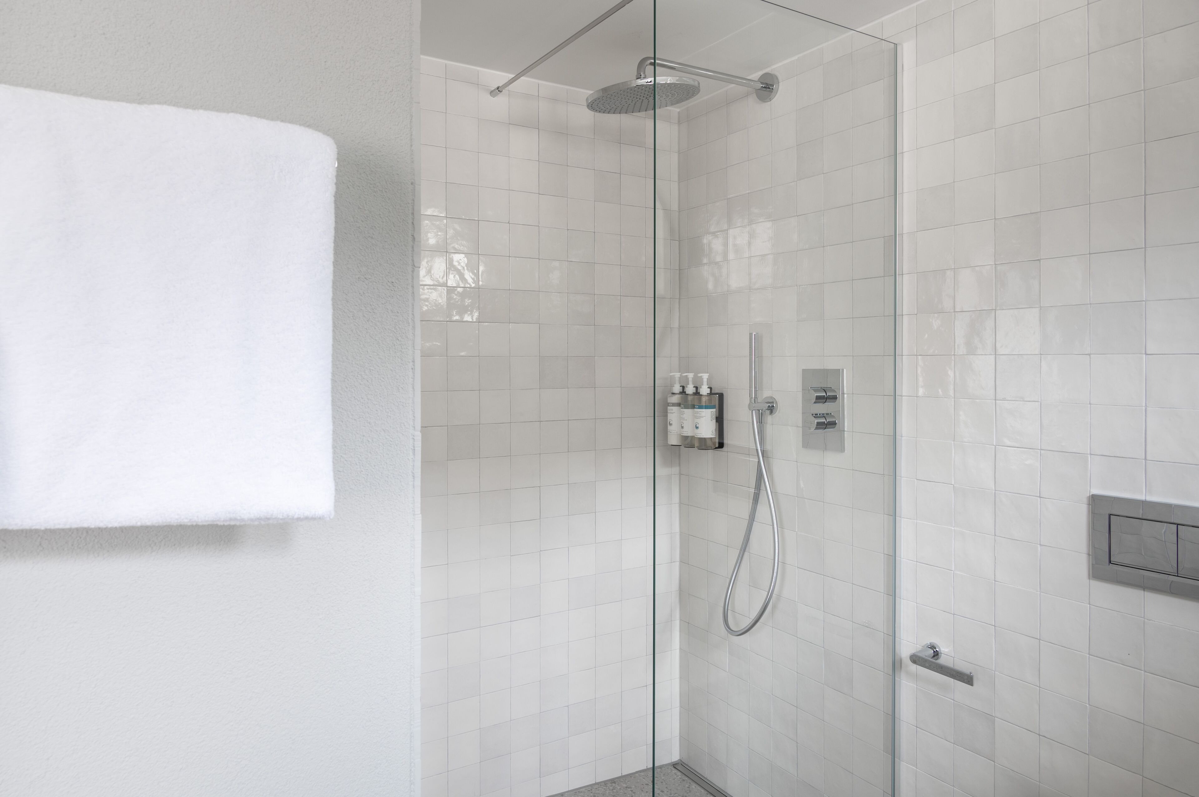 Une douche fermée par des parois de verre, recouverte de carreaux blancs, avec une serviette accrochée au mur et un panneau de commande.