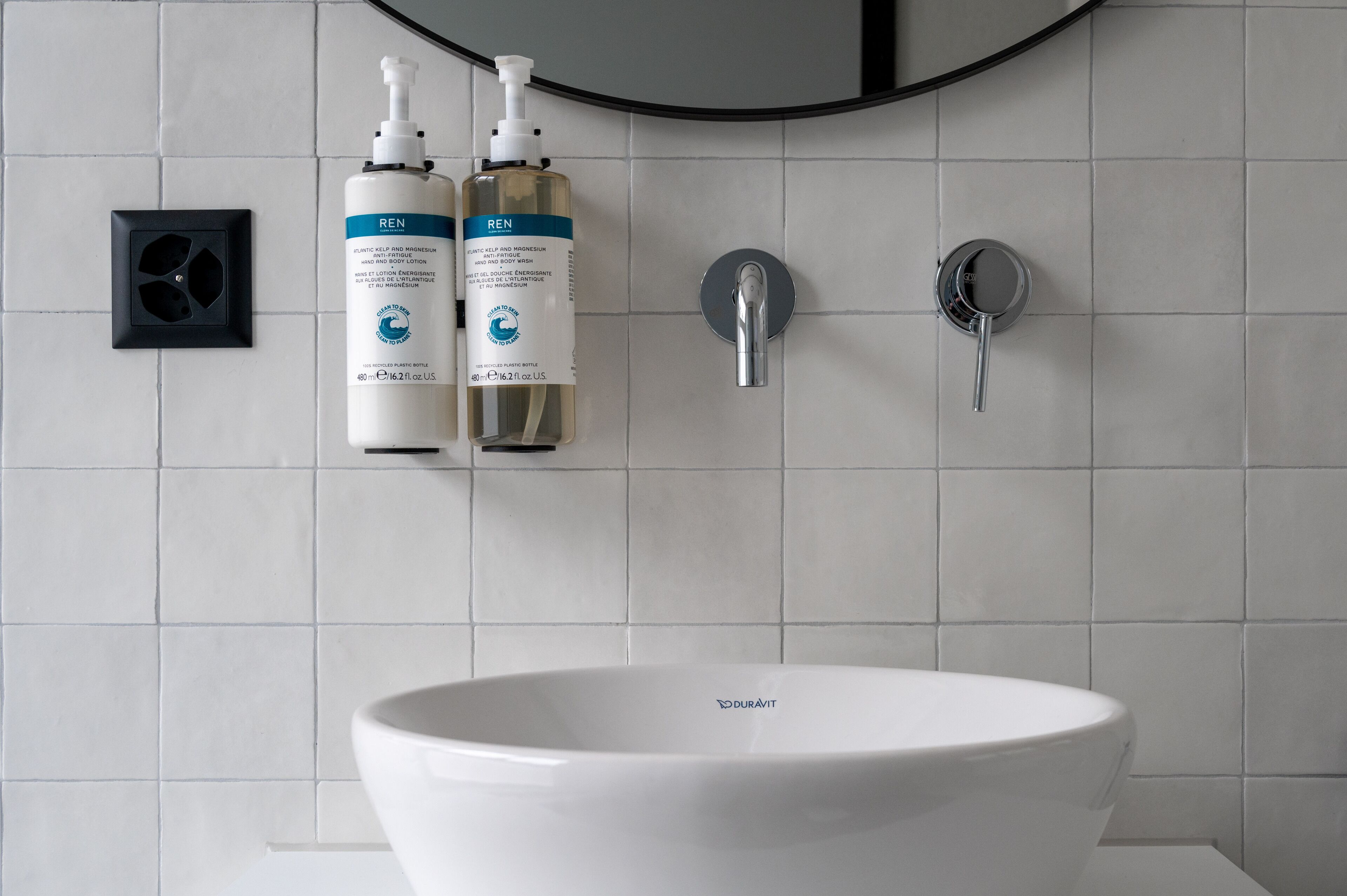 A bathroom with a white sink, two bottles of REN body wash, a mirror, and a showerhead.