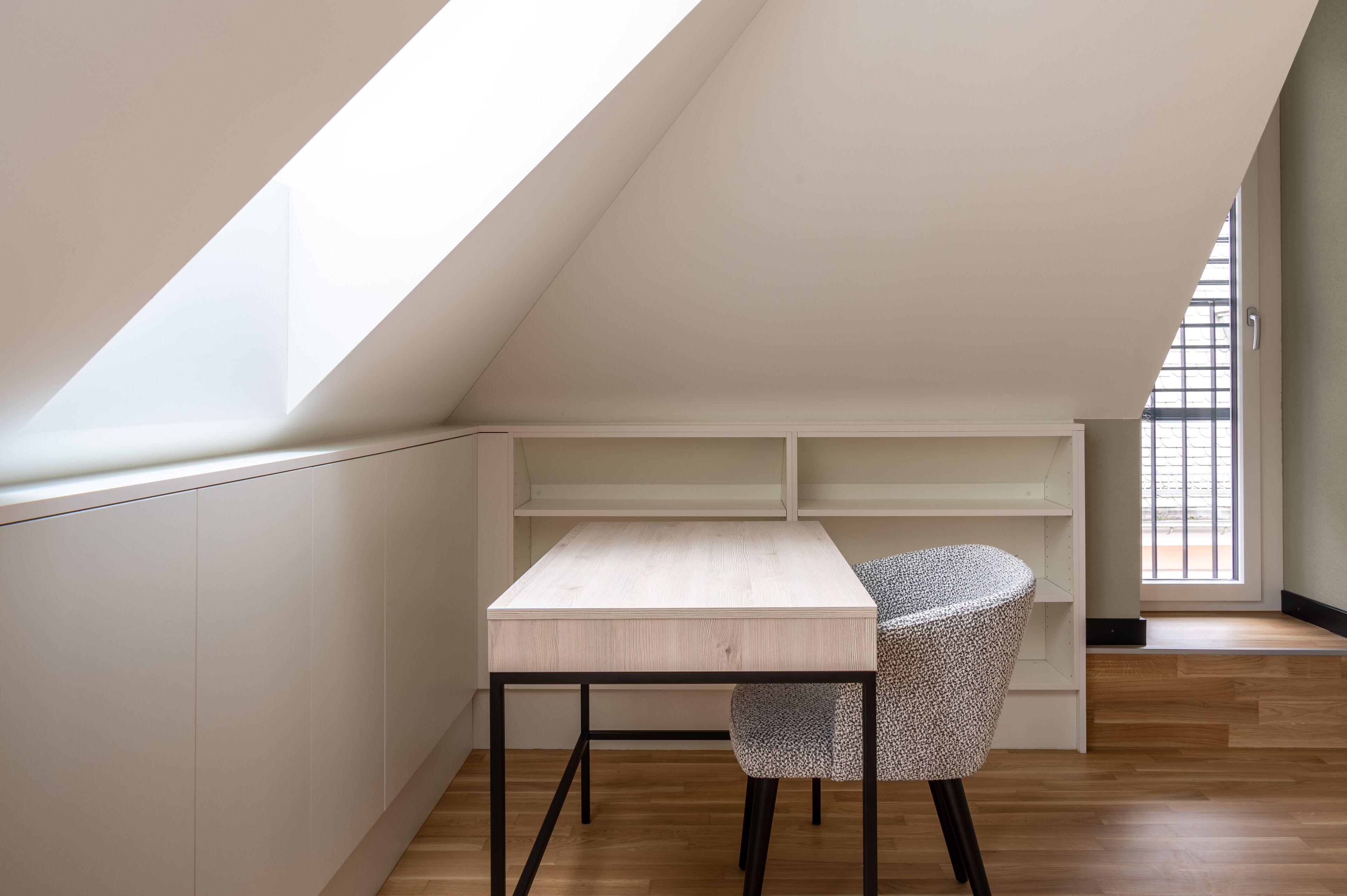 Una habitación pequeña y moderna con techo inclinado, un escritorio de madera con silla y estanterías integradas.