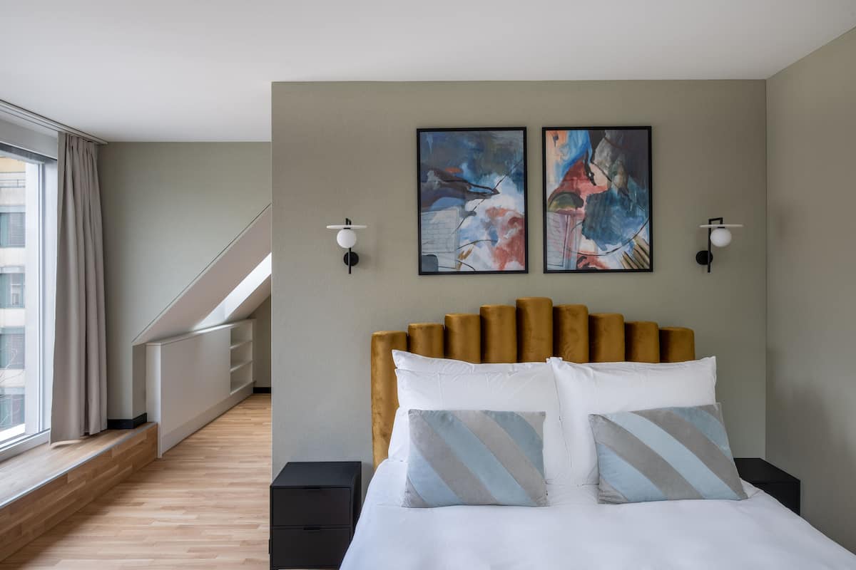 Une chambre d’hôtel moderne avec un grand lit, deux tableaux accrochés au mur, une tête de lit en bois et une vue sur la ville par une grande fenêtre.