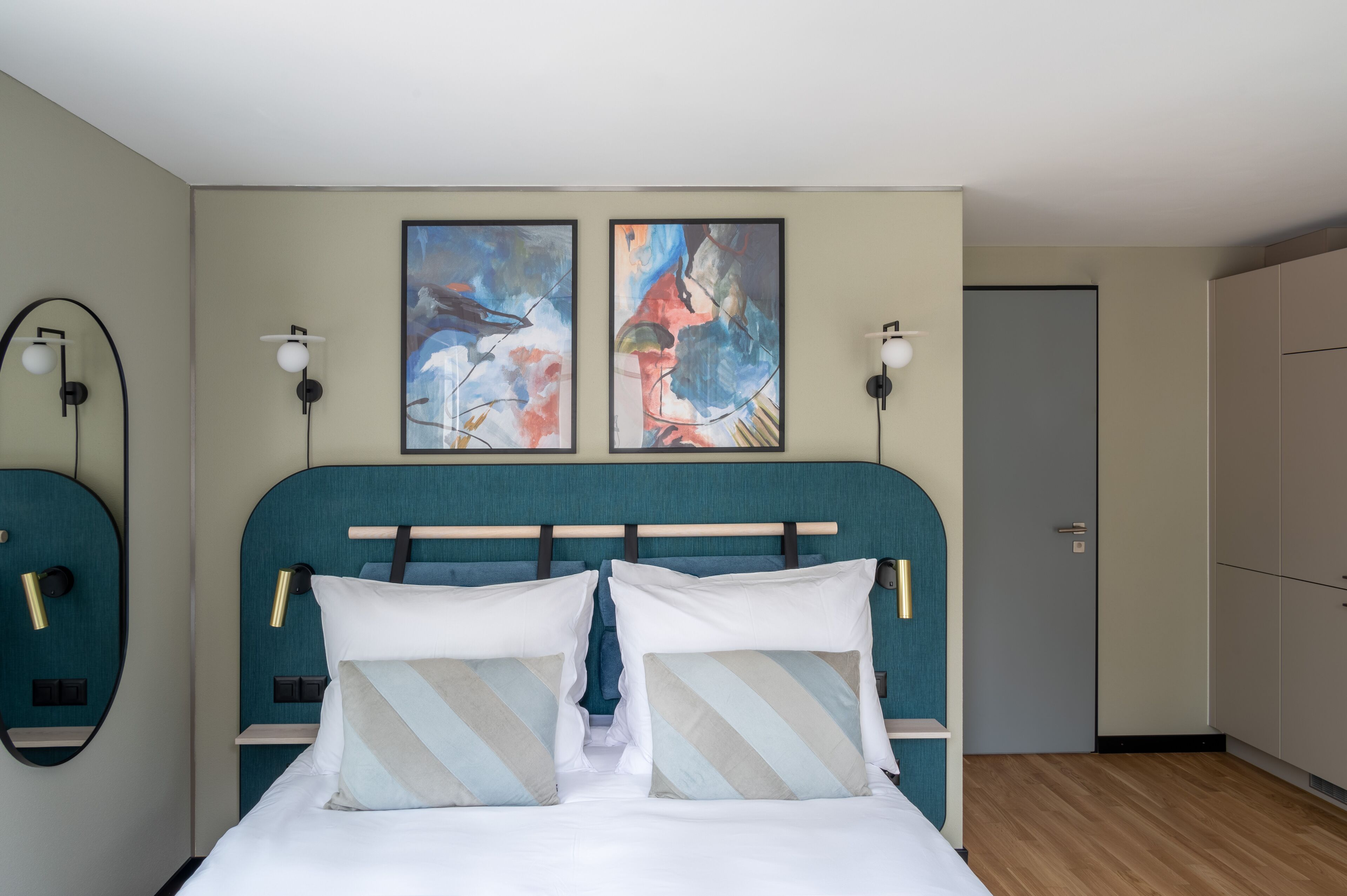 Un dormitorio moderno con un cabecero verde azulado, dos cuadros abstractos, lámparas de noche y un armario.