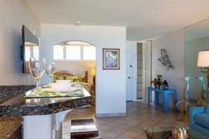 A modern hotel room with a kitchen area, a dining table, and a living area with a sofa and a lamp.