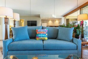 En blå sofa med en farverig pude, et glasterbord og et køkken i baggrunden.