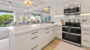 Una cocina moderna con armarios blancos, una isla grande, electrodomésticos de acero inoxidable y vistas a la vegetación exterior.