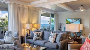 Una sala de estar con un sofá, sillones, una mesa de centro y un televisor. Hay cortinas, un balcón con vistas y un ventilador de techo.