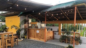 An outdoor seating area with wooden tables and chairs, a bar counter, and various potted plants.