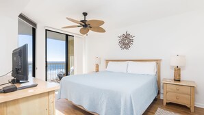 A bedroom with a bed, a desk with a computer, a ceiling fan, a nightstand with a lamp, and a view of the ocean. - Stay @ Folly Ha'i on Folly Beach (Folly Beach)