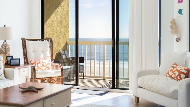 A room with a white sofa, a wicker chair, a wooden table, and a view of the ocean through large glass doors. - Stay @ Folly Ha'i on Folly Beach (Folly Beach)