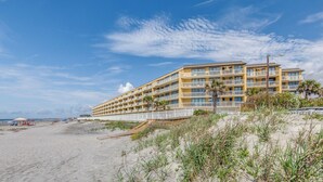 A beachfront hotel with multiple balconies, a sandy beach in the foreground, and a clear sky. - Stay @ Folly Ha'i on Folly Beach (Folly Beach)