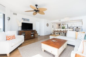 Uma sala de estar espaçosa com um sofá branco, uma mesa de centro de madeira e um ventilador de teto.