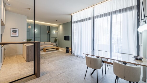 A modern living room with a glass dining table, a sofa, and a kitchen area visible through an open door. - Florella Residences 3 pièces Marceau (Cannes)