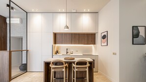 A modern kitchen with a central island, two bar stools, pendant lighting, and a framed artwork on the wall. - Florella Residences 3 pièces Marceau (Cannes)