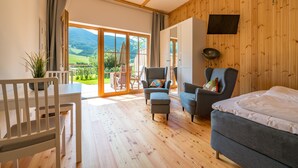Una habitación amplia con suelo de madera, una cama, dos sillones, una mesa de comedor y un ventanal que ofrece vistas a un paisaje verde.