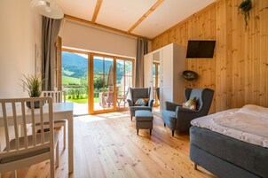 Um quarto espaçoso com piso de madeira, uma cama, duas poltronas, uma mesa de jantar e uma grande janela com vista para uma paisagem verde.
