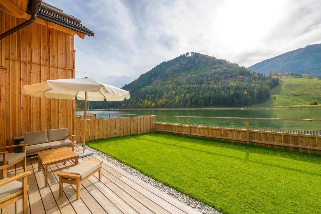 A wooden deck with outdoor furniture, a white umbrella, and a view of a lake and mountains.