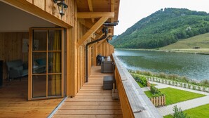 Una terraza de madera con puerta de vidrio que da a una habitación, con vistas a un lago y colinas.