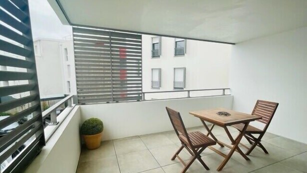 En balkon med et bord og stole, en plantekasse og udsigt til bygninger gennem metaljalousier.