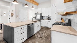 Una cocina moderna con armarios blancos, una isla central, electrodomésticos de acero inoxidable, iluminación colgante y una viga de techo de madera.