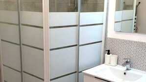 Een badkamer met een douchecabine van glas, een witte wastafel met spiegel, en een betegelde spatwand.