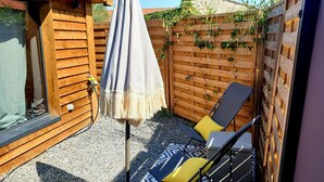 Een houten terras met een parasol, twee lounge fauteuils en een houten omheining begroeid met klimplanten.