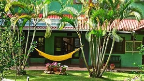 Una casa verda amb teulada vermella, envoltada de vegetació abundant i una hamaca.