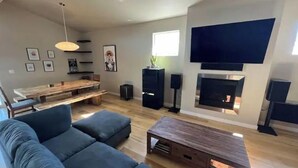 Ein modernes Wohnzimmer mit einem Ecksofa, einem Holztisch, einem Kamin, einem an der Wand montierten Fernseher und einem Essbereich mit Holztisch und Stühlen.