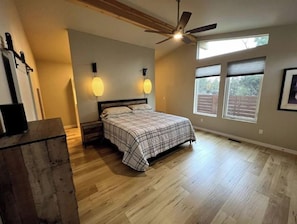 A bedroom with a bed, wooden floor, ceiling fan, two wall-mounted lights, and windows.