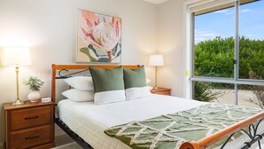 A bedroom with a bed, two bedside tables, a nightstand, a window with a view of greenery, and a painting on the wall.