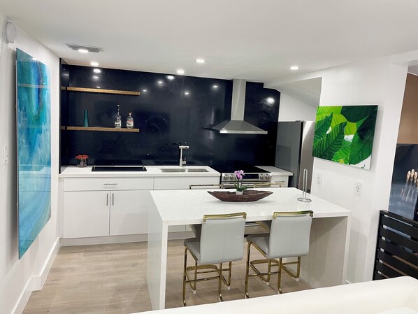 Een moderne keuken met witte kastjes, een zwarte achterwand en een rvs afzuigkap. Er is een eethoek met een witte tafel en twee stoelen.