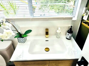 Een badkamer met een witte wastafel, een gouden kraan, een kleine potplant en een handzeepdispenser.