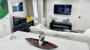 Un salon moderne avec un canapé blanc, une table basse en bois ornée d’un vase de fleurs, un meuble TV noir avec un téléviseur à écran plat et une fenêtre avec des rideaux.