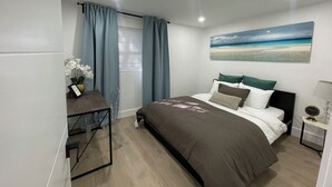 Une chambre à coucher avec un lit, un bureau avec une chaise, une fenêtre avec des rideaux, une table de chevet avec une lampe et un tableau représentant une plage.