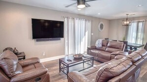 Una sala de estar con sofás de cuero, un televisor de pantalla plana, un ventilador de techo y un comedor con mesa y sillas.