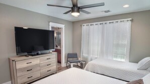 Un dormitorio con dos camas, televisión, cómoda, ventilador de techo y una ventana con cortinas.