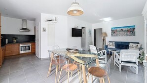 A modern kitchen with white cabinets, a built-in oven, and a black countertop. There is a dining area with a glass table and chairs, and a blue sofa with decorative pillows. - VILLA SOLEIL (La Brée-les-Bains)