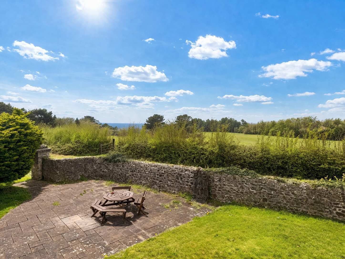 A stone wall, a paved patio, and a view of a green field and trees.