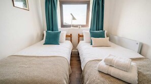 A twin bedroom with two beds, teal curtains, a window with a view, and a bedside table with a lamp.