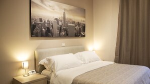 Ein Hotelzimmer mit Bett, Nachttisch mit Lampe, Stadtbildgemälde an der Wand und einem Fenster mit Vorhängen.