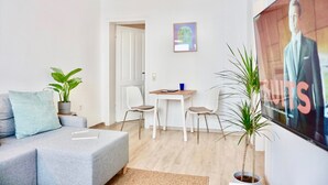 Een moderne woonkamer met een grijze bank, twee potplanten, een kleine tafel met stoelen en een lijst met een foto aan de muur.